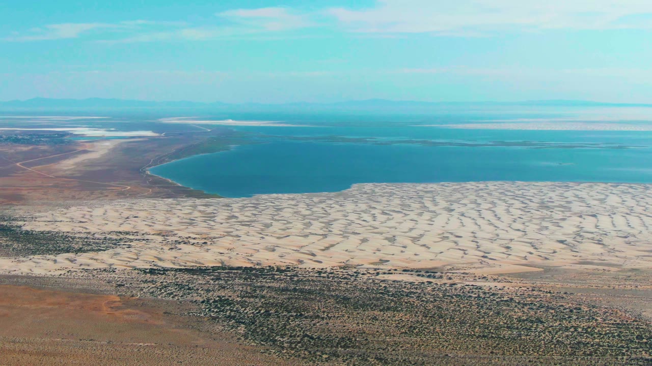 Aerial view in Guerrero Negro where pale sandy shoreline forms a bright band between blue water and sparse desert vegetation, no visible people.