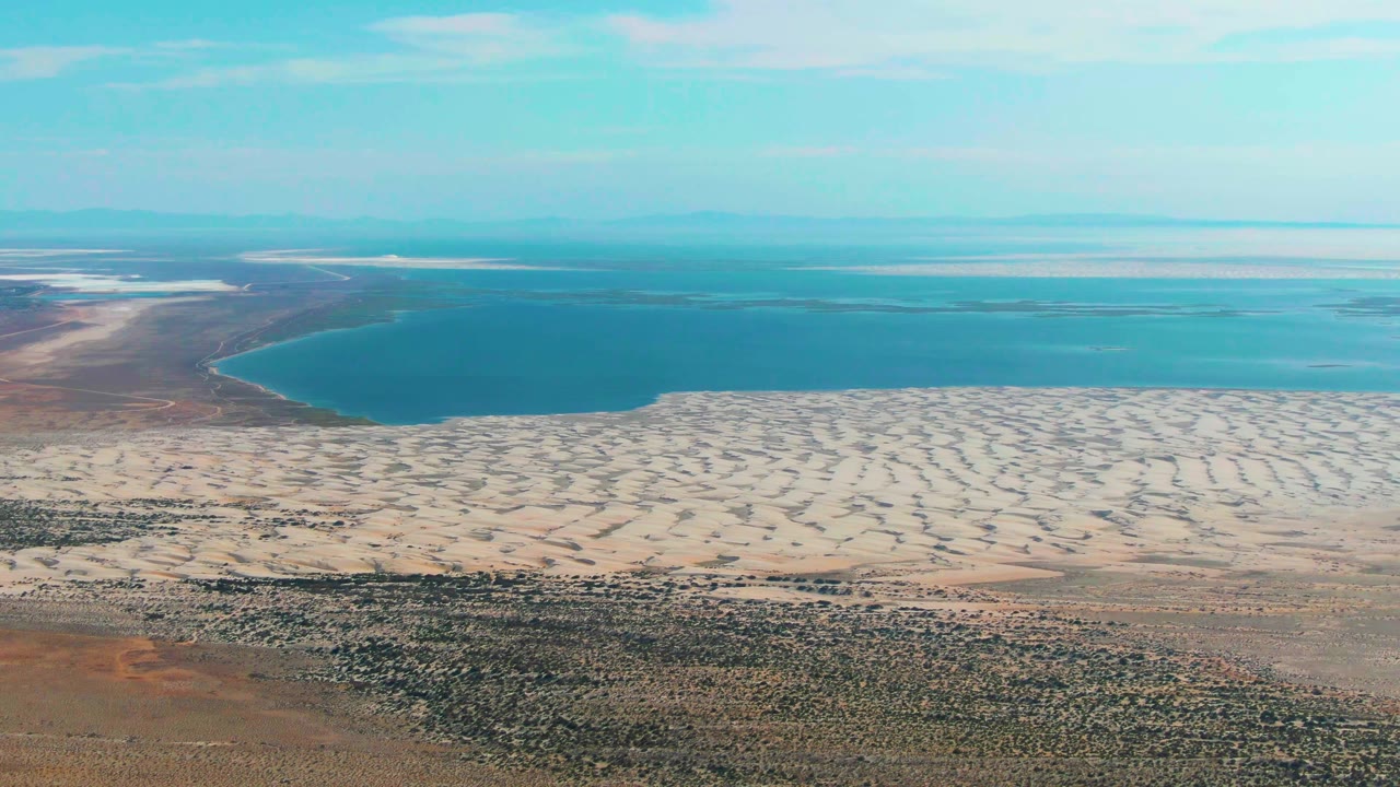 Wide aerial view of a blue lake, pale dunes, scrubland, and light blue sky.