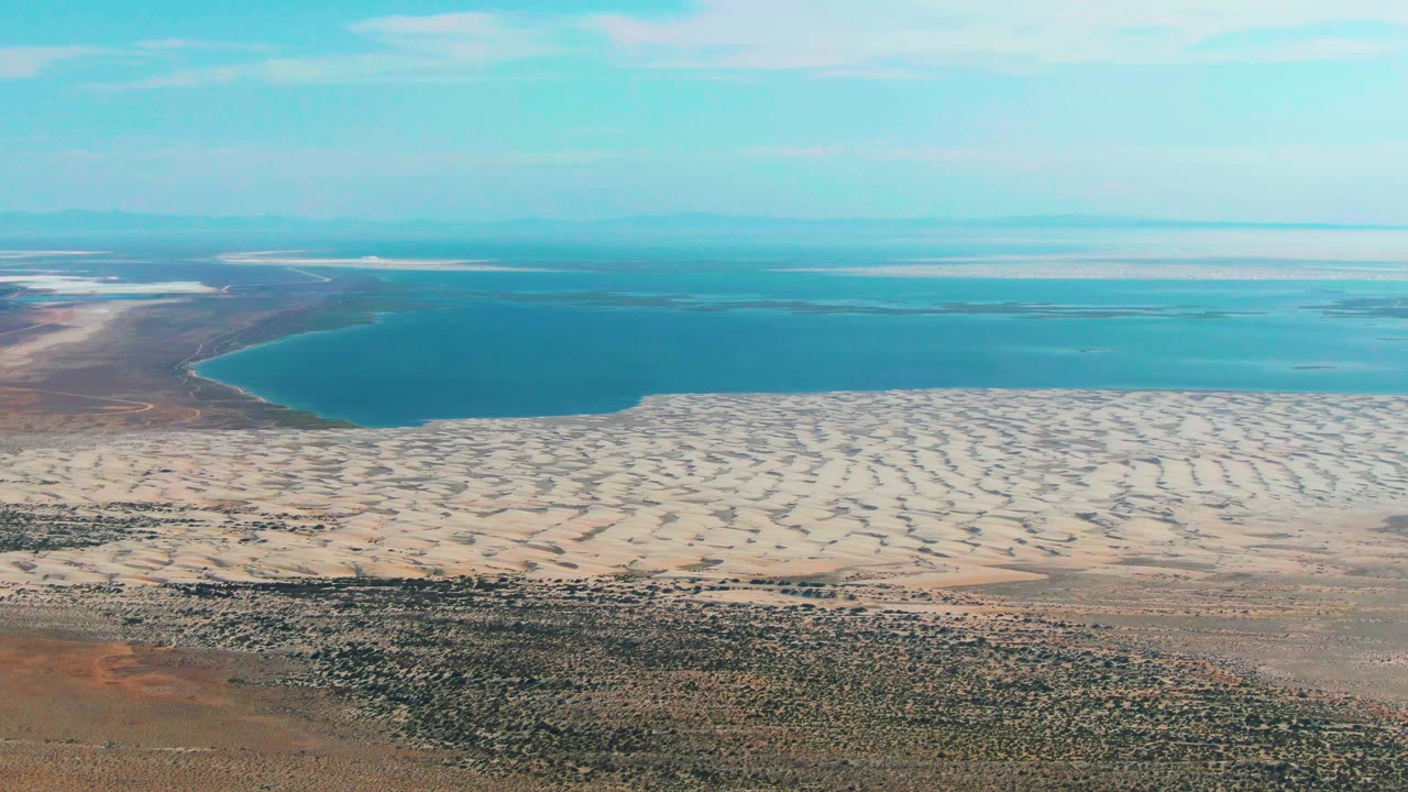 Second aerial frame in Guerrero Negro shows the blue lake slightly left of center, a wide pale dune field below it, distant water bands, and no visible people.