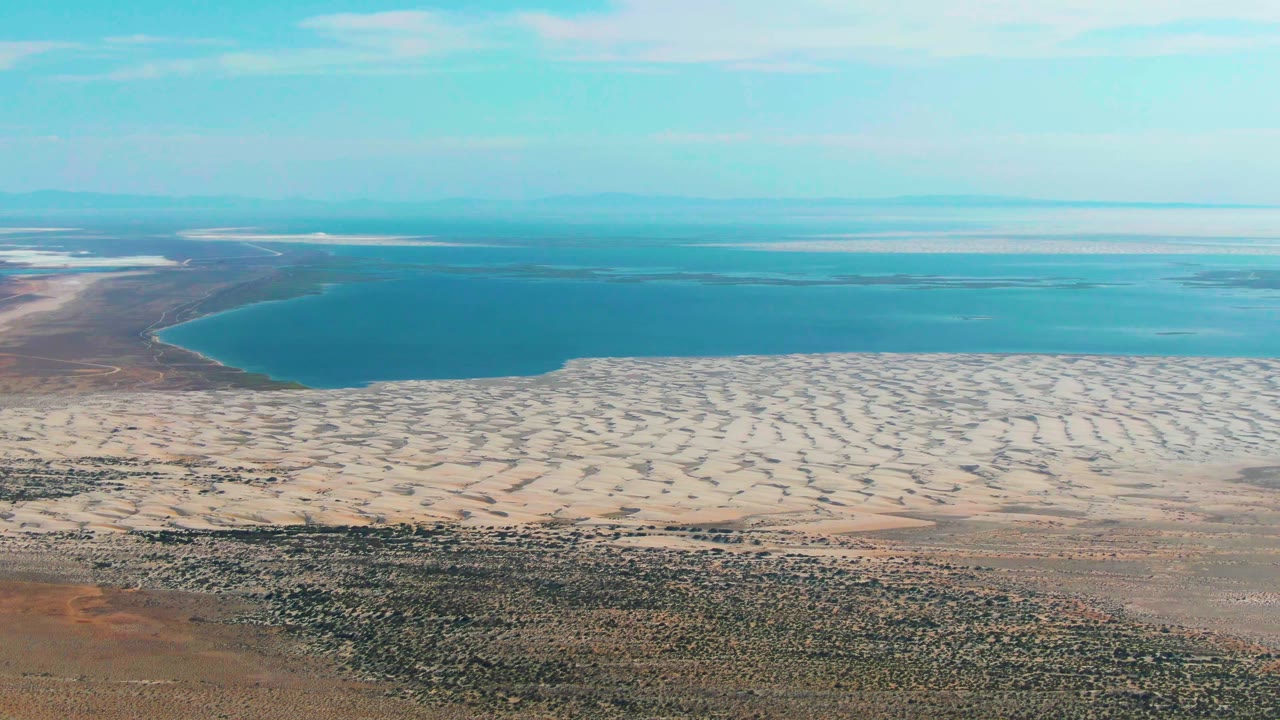 Third frame presents a high aerial view over Guerrero Negro with dark scrubland in front, pale sand ridges through the middle, the blue lake beyond, and no visible people.