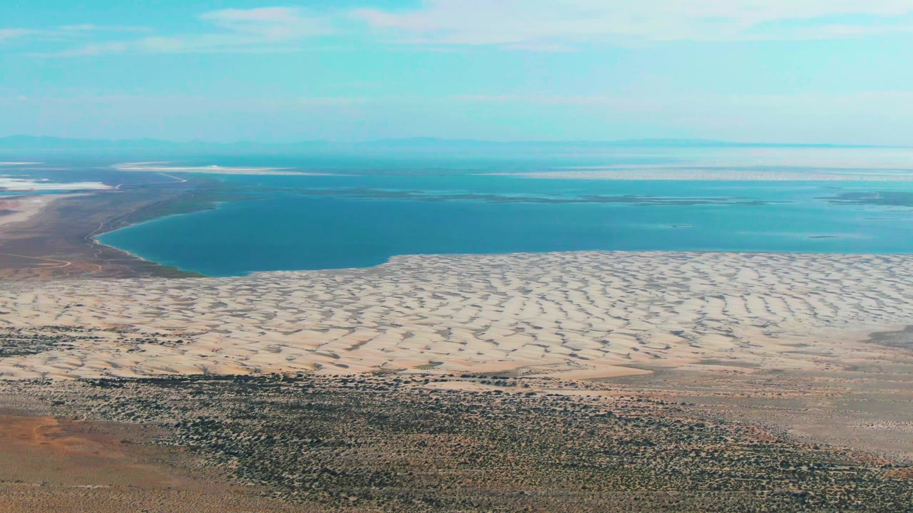 Fourth frame in Guerrero Negro holds on curved blue water behind a broad pale sand field, with darker dry scrub across the lower frame and no visible people.