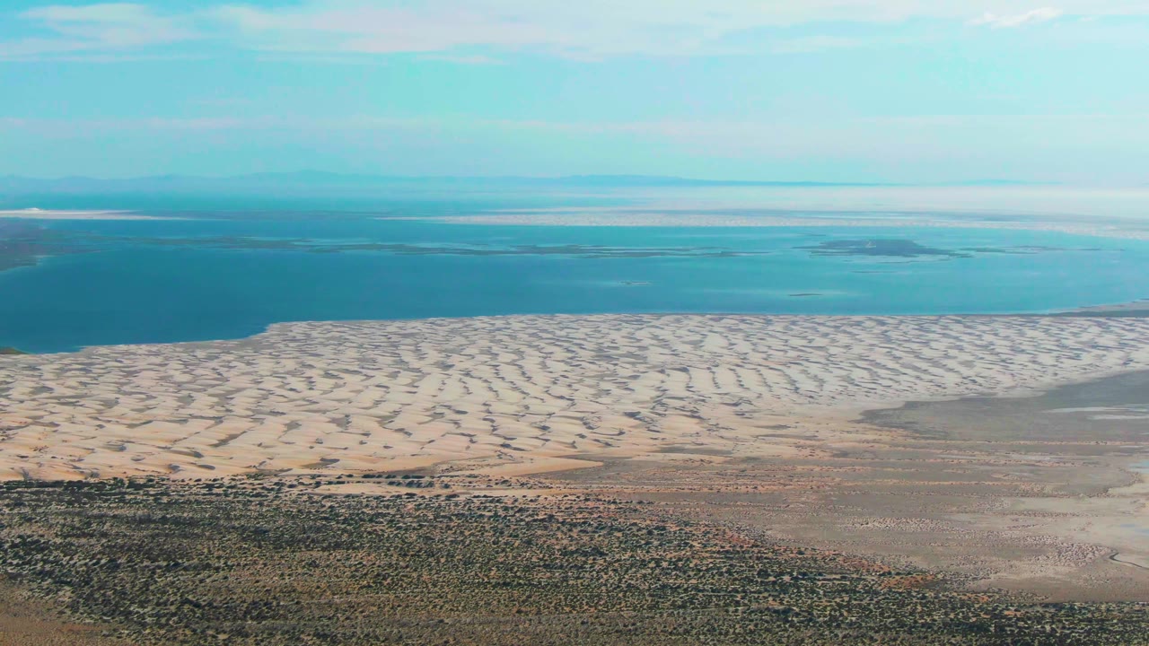 Aerial frame over Guerrero Negro, Baja California Sur, showing pale rippled tidal sand, blue coastal water, scrubby foreground land, and no visible people.
