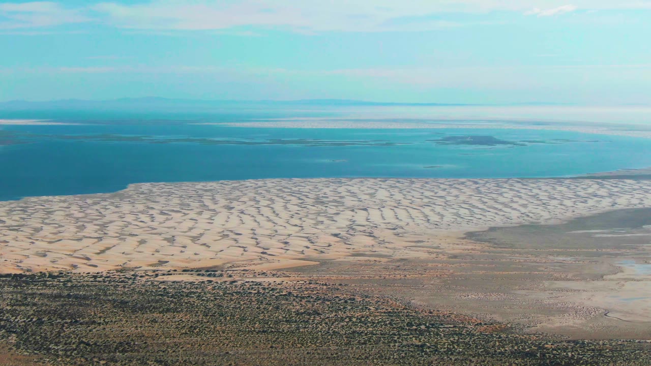 Aerial Guerrero Negro coastline frame with pale ripple marks in the sand, calm blue water beyond, darker dry land at the bottom edge, and no visible people.