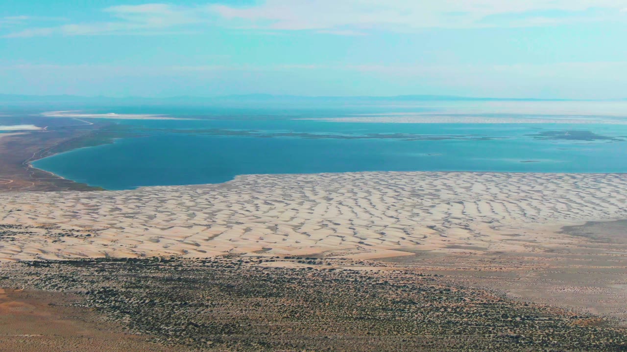 Aerial frame over Guerrero Negro, Baja California Sur, showing dry foreground terrain, pale patterned sand, blue water, distant horizon, and no visible people.