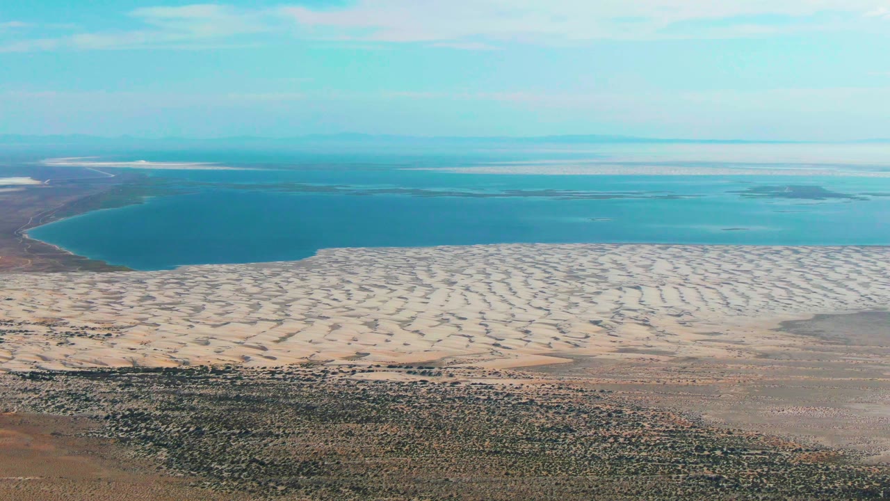 Aerial view in Guerrero Negro with a curved blue water edge, rippled pale sand across the midground, dry desert foreground, and no visible people.