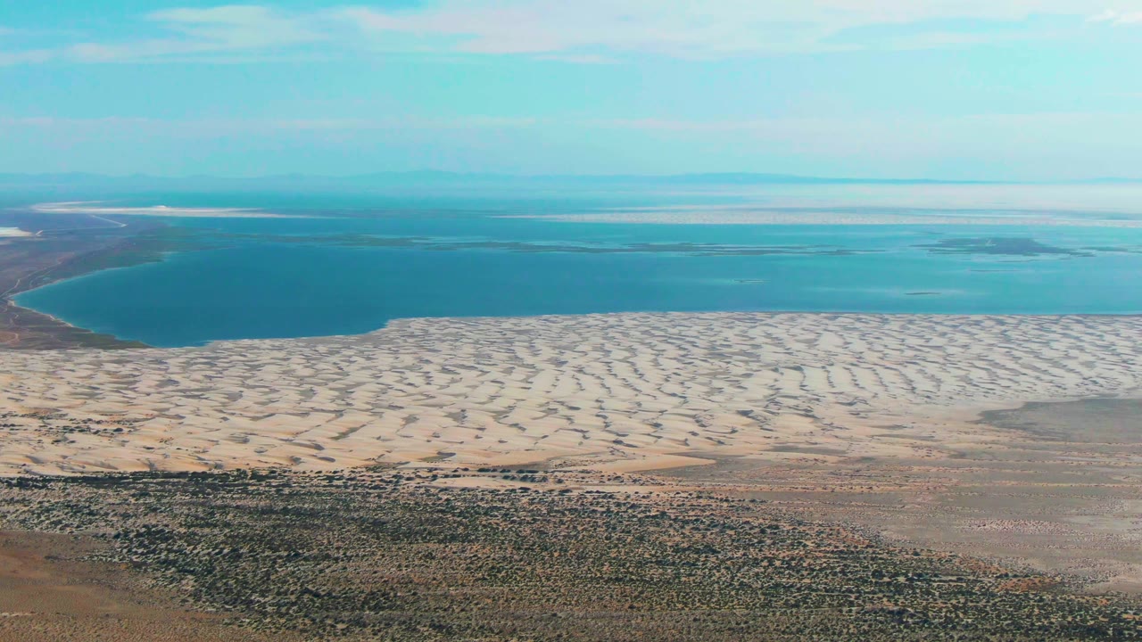 Wide aerial frame over Guerrero Negro showing patterned sand between the rocky foreground and blue water, with a hazy horizon and no visible people.