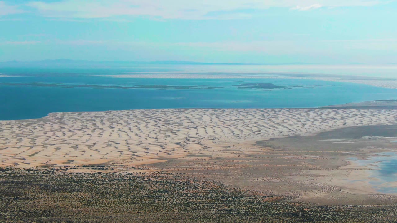 Opening aerial frame over Guerrero Negro, Baja California Sur, with scrubland, pale rippled dunes, shallow flats, blue ocean, a distant landmass, and no visible people.