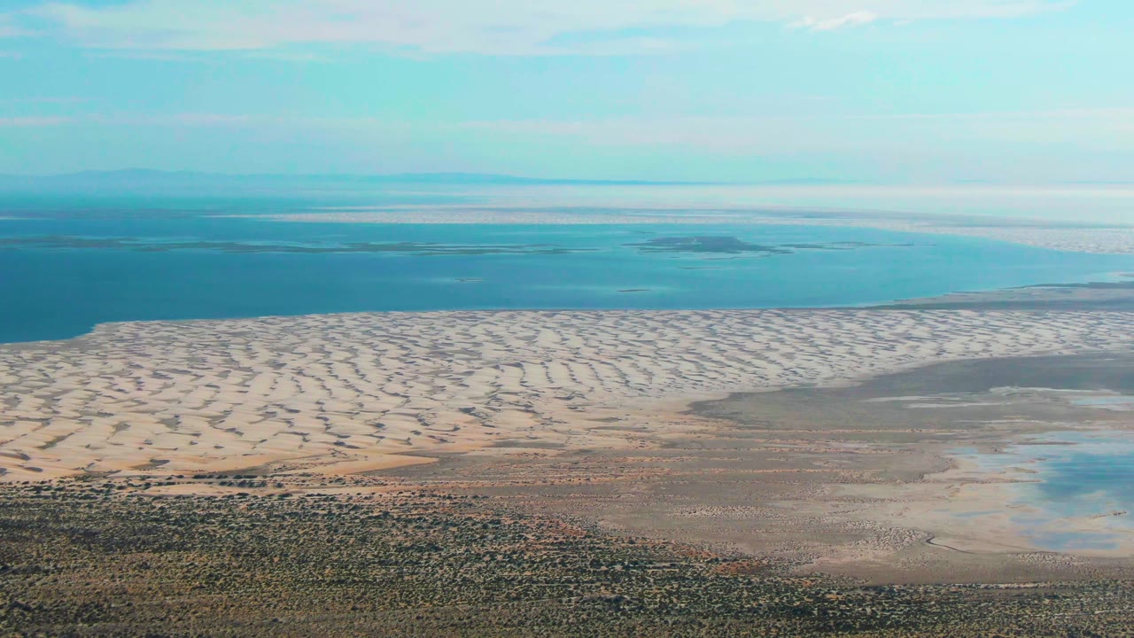 Third frame over Guerrero Negro with patterned pale dunes in the lower middle, brown-gray tidal flats to the right, blue ocean across the background, and no visible people.
