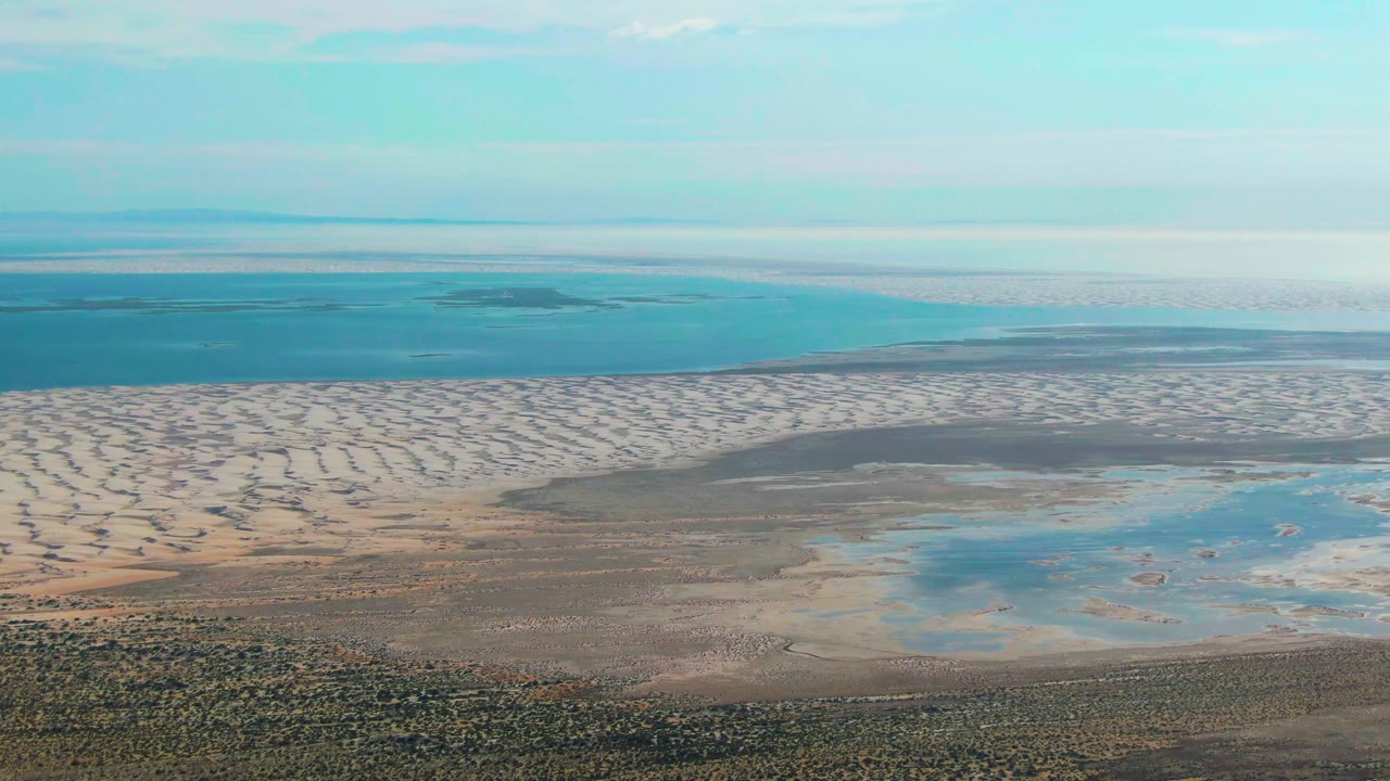 Opening aerial frame over Guerrero Negro, Baja California Sur, with pale patterned tidal flats, blue pools, distant ocean, light clouds, and no visible people.