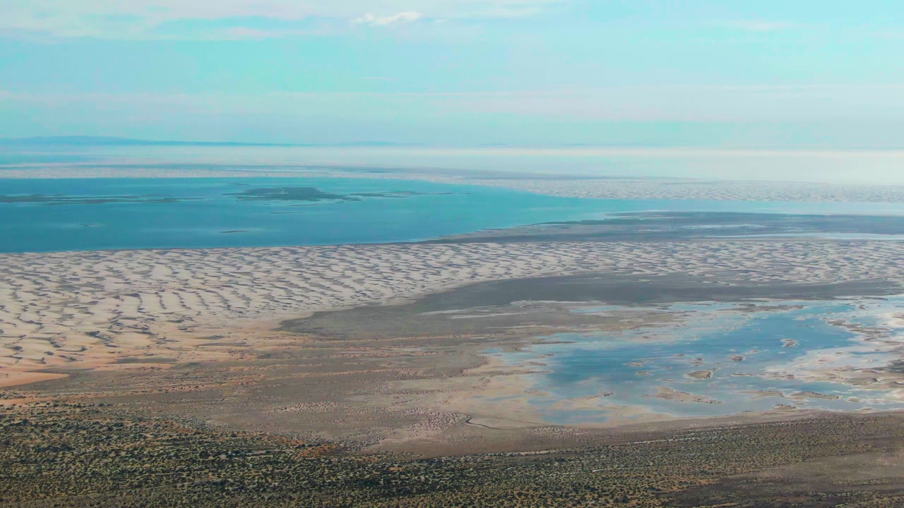 Guerrero Negro aerial frame with sandy patterned flats stretching toward the ocean, shallow blue water in the lower right, hazy horizon, and no visible people.