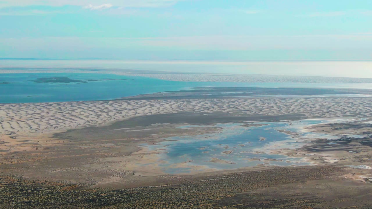 High aerial view over Guerrero Negro tidal flats with pale sandbars, shallow blue pools, calm ocean beyond, and no visible people.