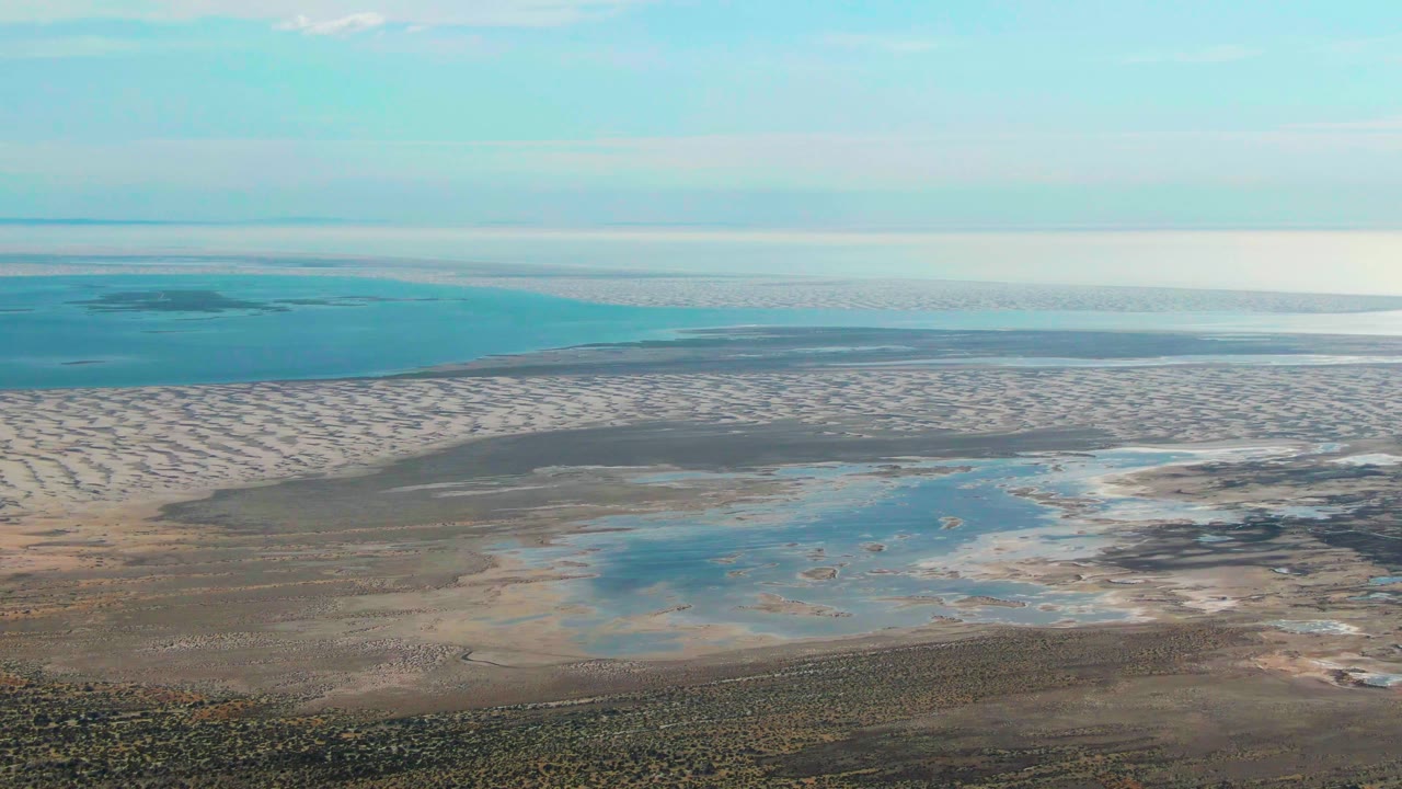 Aerial frame over Guerrero Negro shoreline showing wet sand patterns, a central blue water pool, distant ocean, clear sky, and no visible people.
