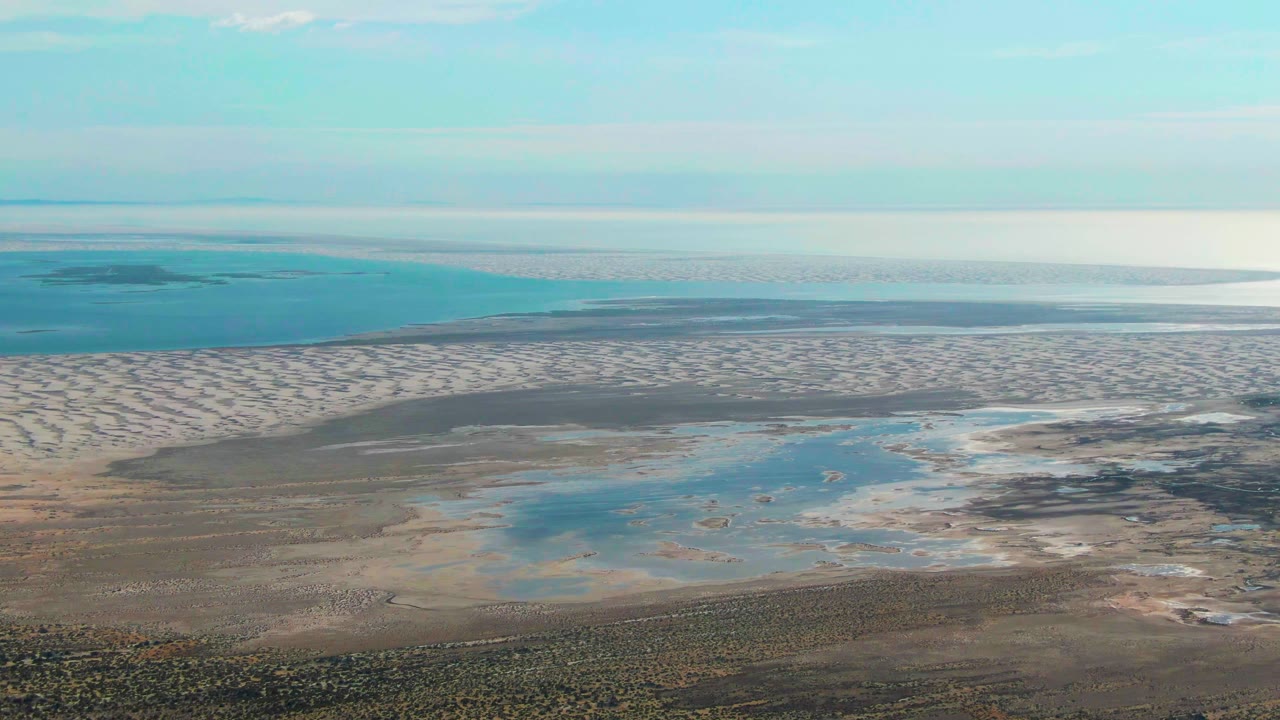 High oblique view of Guerrero Negro tidal sand with shallow blue water, pale exposed flats, darker wet patches, low ocean horizon, and no visible people.
