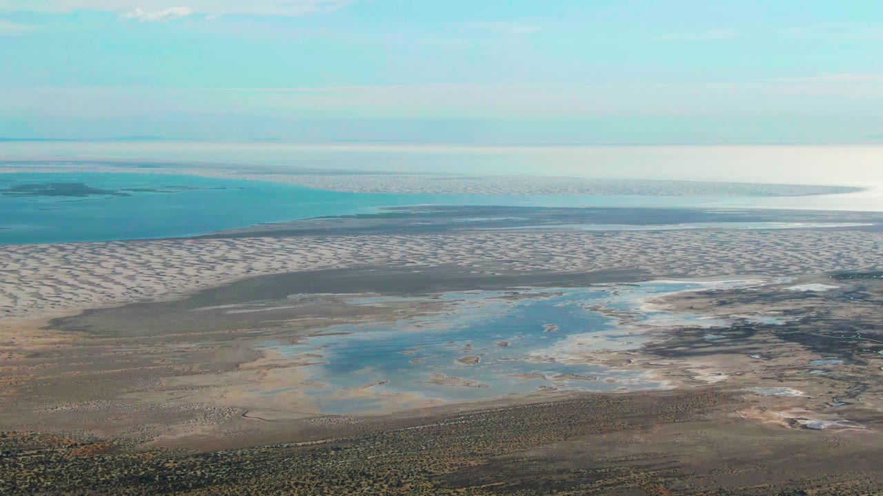 Later aerial frame over Guerrero Negro with exposed sandbars across the midground, shallow pools near the shore, calm sea in the distance, and no visible people.