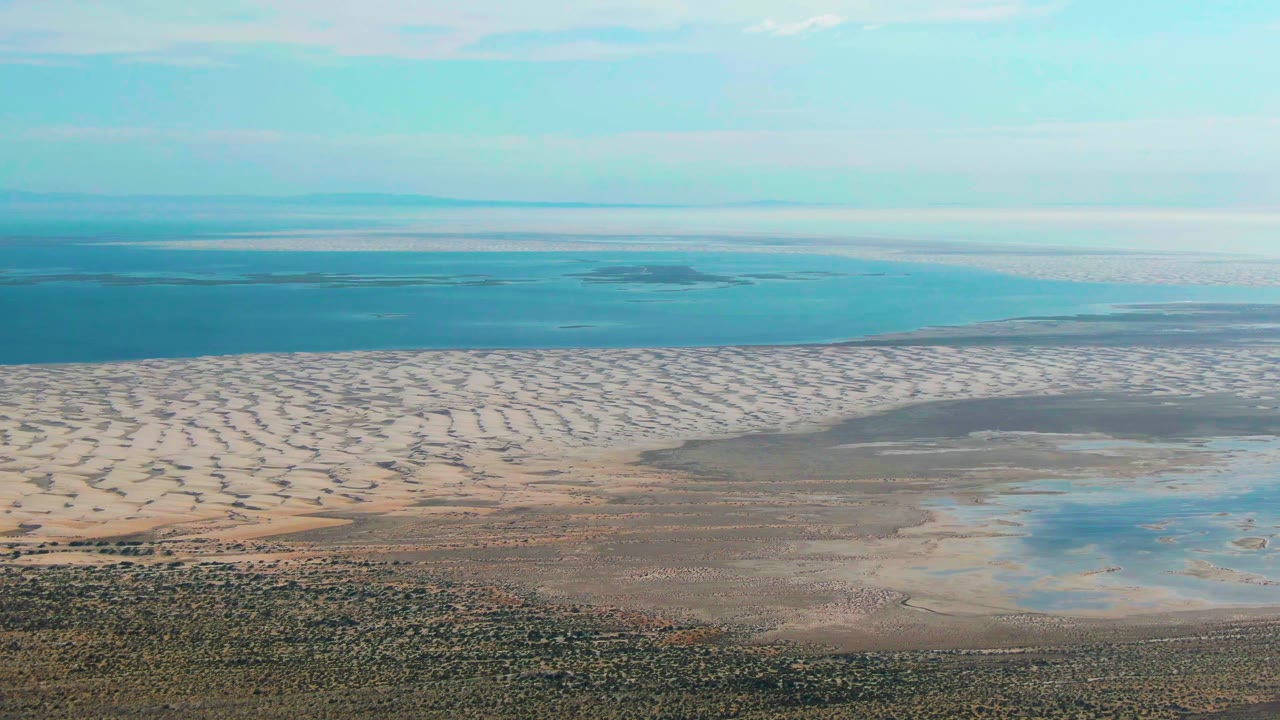 Aerial view over Guerrero Negro showing scrubby foreground, pale coastal dunes, wetland pools, blue ocean, thin clouds, and no visible people at the start of the clip.