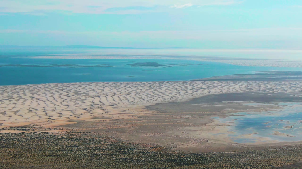 Wide aerial frame over Guerrero Negro with pale dunes across the middle, blue ocean beyond, reflective wetland water to the lower right, and no visible people.