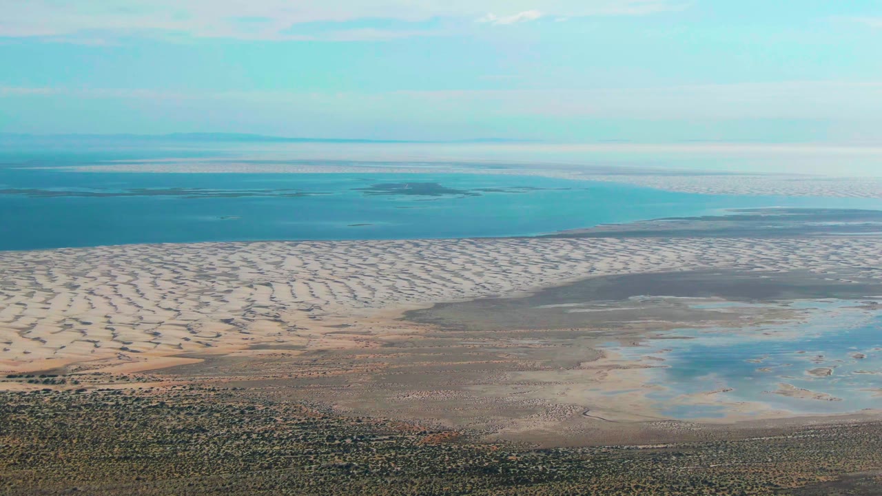 Aerial view in Guerrero Negro with a broad band of light dunes, shallow lagoon water on the right, darker ocean on the left horizon, and no visible people.