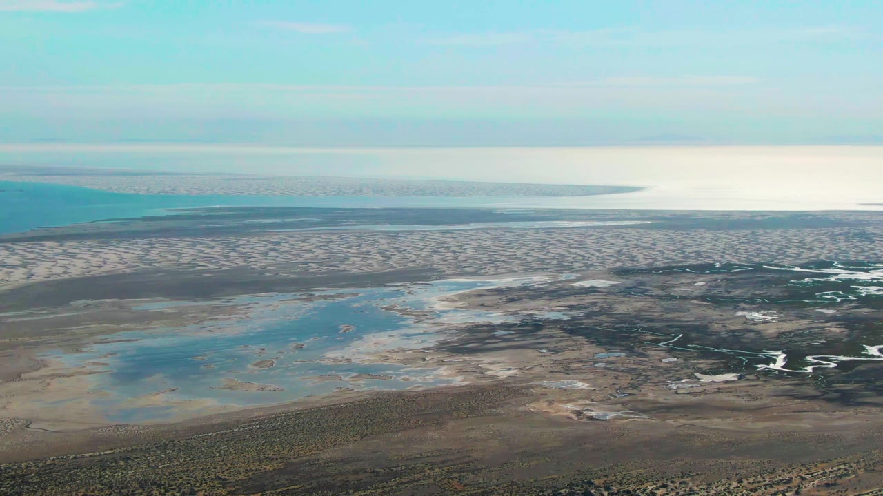 Aerial view in Guerrero Negro, Baja California Sur, Mexico showing sandy tidal flats, shallow blue pools, darker wet channels, distant ocean, clear sky, and no visible people at the start of the clip.