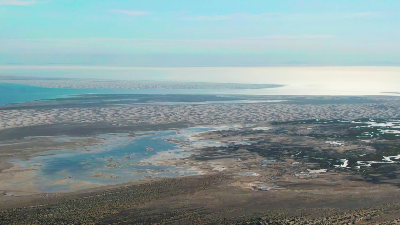 High aerial frame over Guerrero Negro tidal shoreline with pale sandbars, turquoise pools, dark wet sand, low scrub at the bottom edge, distant ocean water, clear sky, and no visible people.
