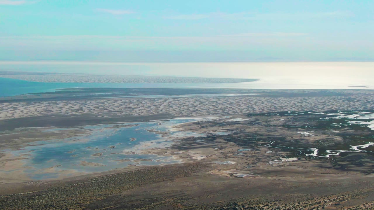 Wide aerial view in Guerrero Negro, Mexico with lagoon-like blue pools in the sand, branching dark tidal channels to the right, dry scrubland in the foreground, bright ocean horizon, and no visible people.