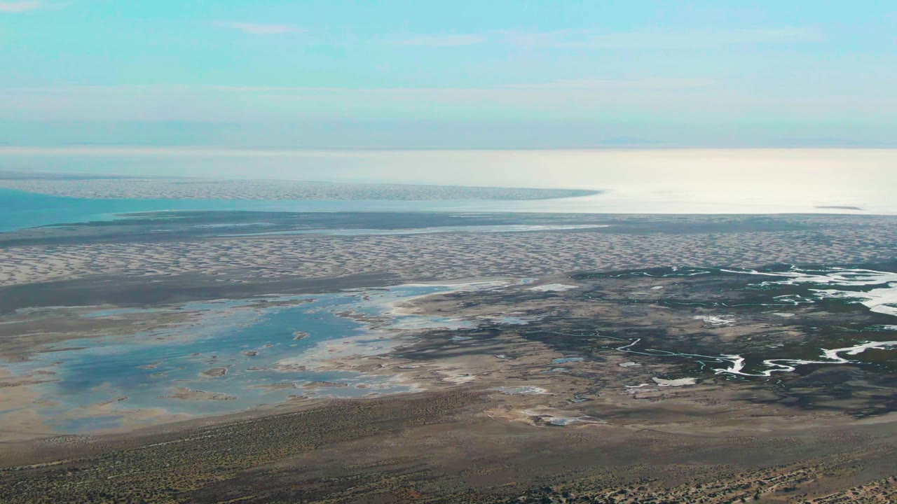 Aerial coastal flats with shallow blue pools, pale sand, and distant waterline.