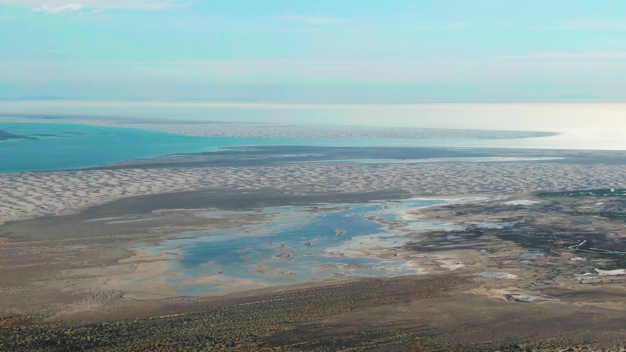 Aerial frame over Guerrero Negro tidal flats with turquoise pools, beige wet sand, ocean on the horizon, scrubland at the bottom edge, and no visible people.