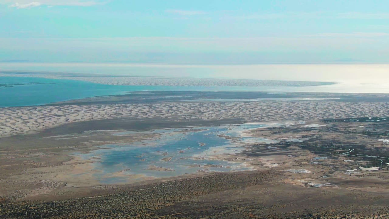 Guerrero Negro aerial view with patterned tidal sand, several pale blue pools, darker wet channels, distant ocean, hazy horizon, and no visible people.