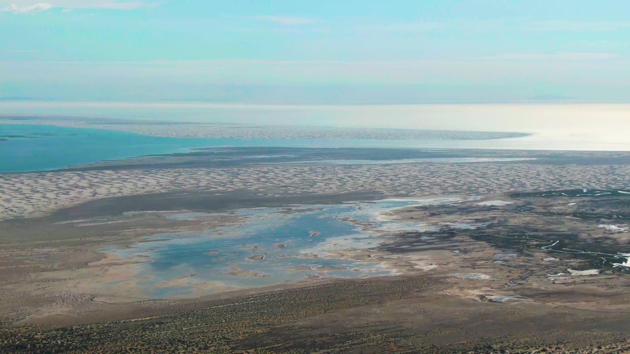 High aerial frame of Guerrero Negro shoreline with a broad band of tidal flats, scattered pools in the midground, ocean and sky beyond, and no visible people.