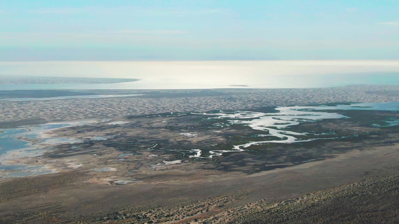 Wide aerial frame over Guerrero Negro, Baja California Sur, showing bright winding water channels across muddy wetland flats, scrub in the lower foreground, and no visible people.