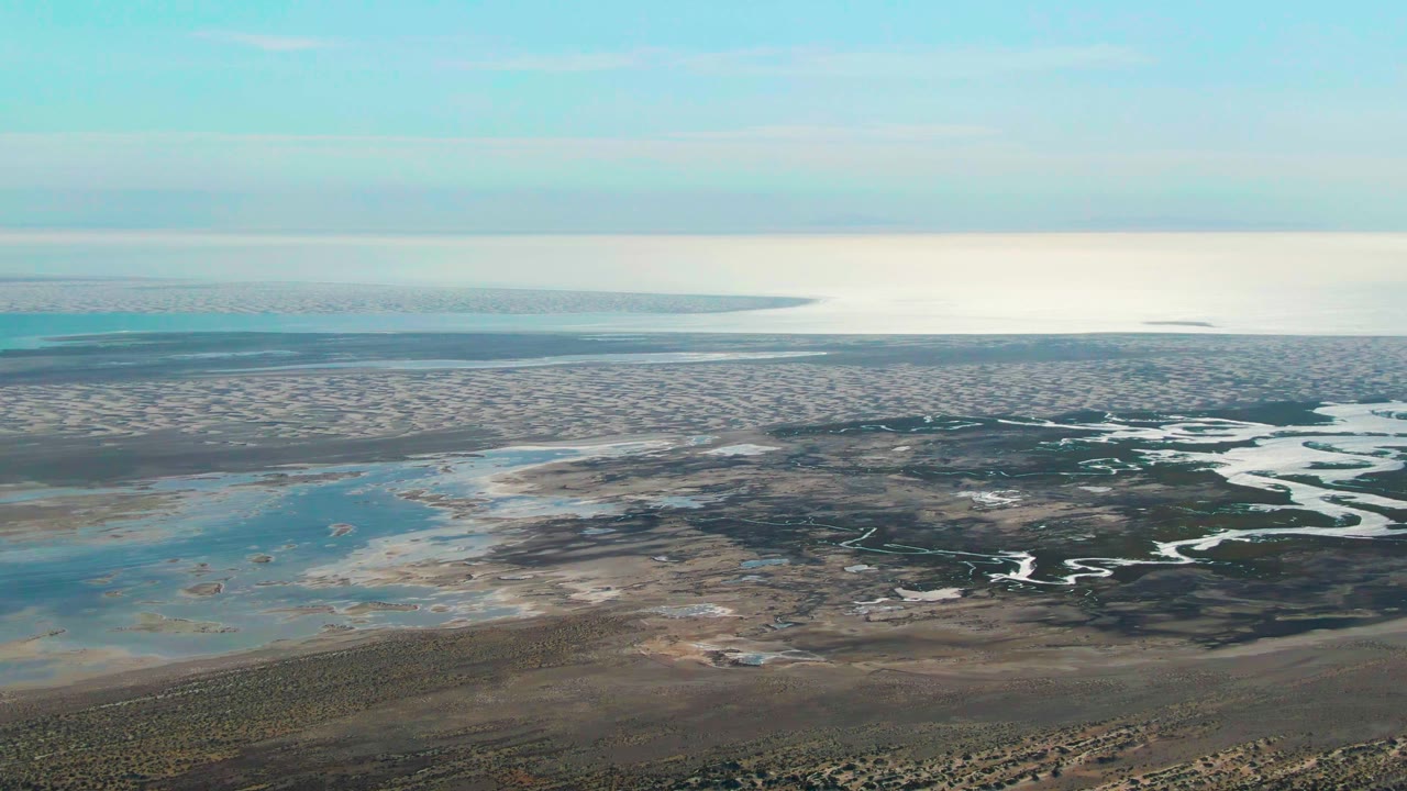 High aerial frame of Guerrero Negro wetlands showing pale shallow water on the left, dark marsh channels through the middle, a flat water horizon, and no visible people.