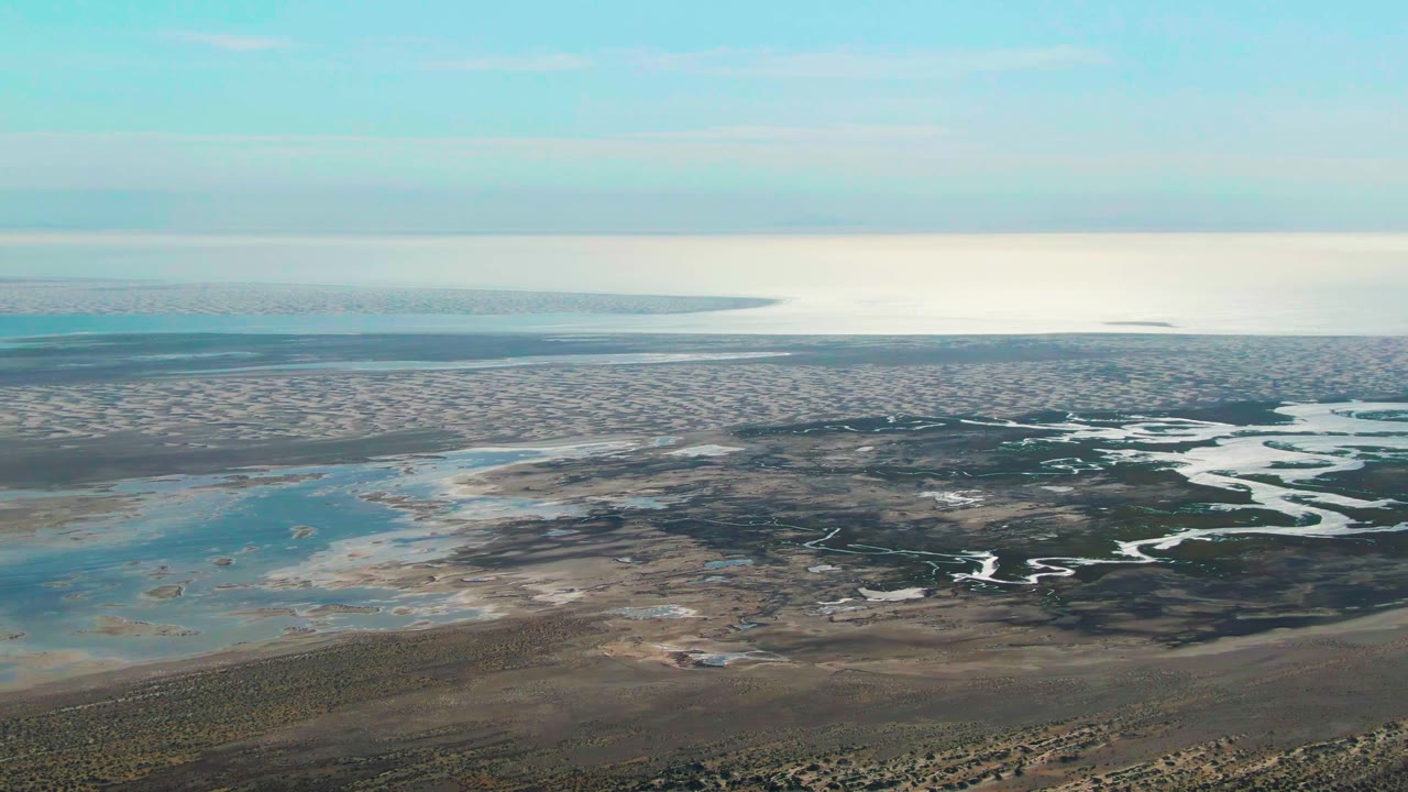 Wide aerial view in Guerrero Negro with a bright horizon above layered mudflats, shallow water pools, dark marsh textures, and no visible people.