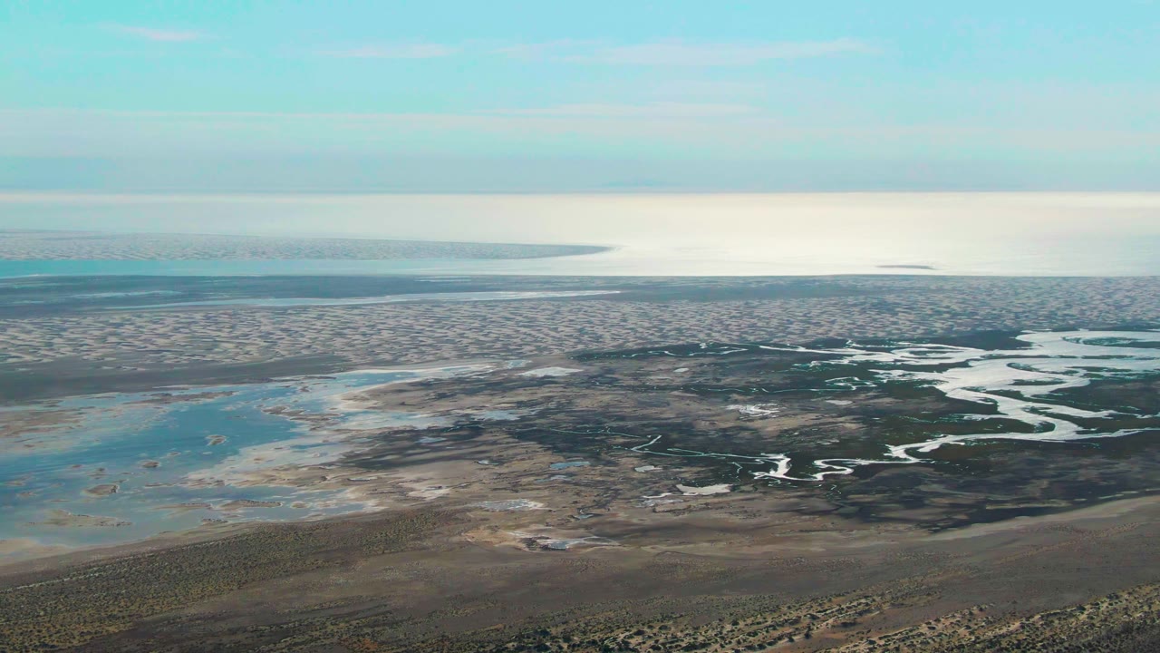 Aerial Guerrero Negro wetland frame with winding channels cutting across brown mudflats, pale blue water to the left, a soft cloud band above, and no visible people.