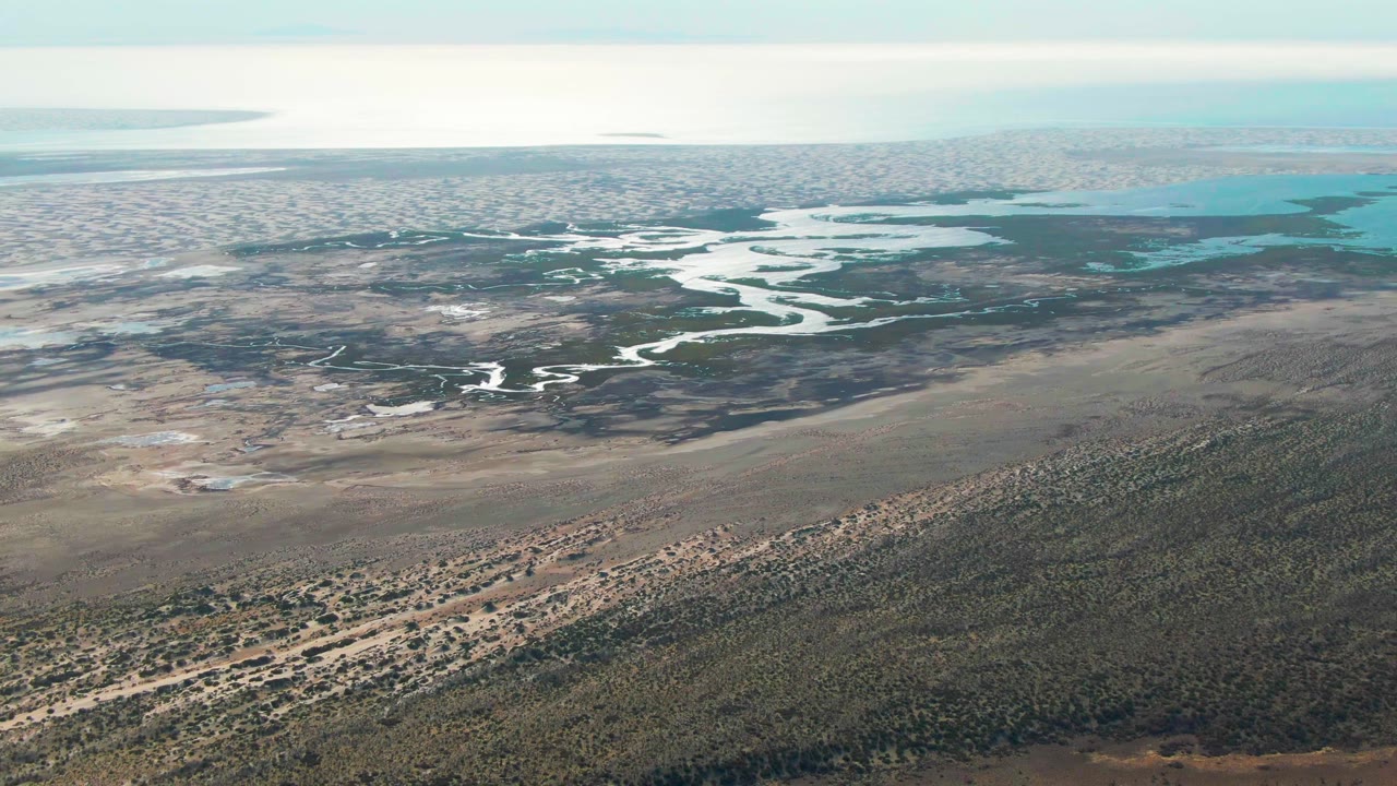Opening aerial frame over Guerrero Negro wetlands with brown dry ground in front, dark green vegetation through the center, pale winding water channels, turquoise pools near the horizon, and no visible people.