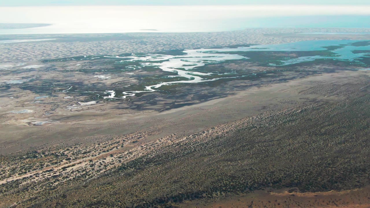 Mid-clip aerial frame over Guerrero Negro shows the wetland channels more centered, with brown flats below, dark green vegetation across the middle, turquoise water beyond, and no visible people.
