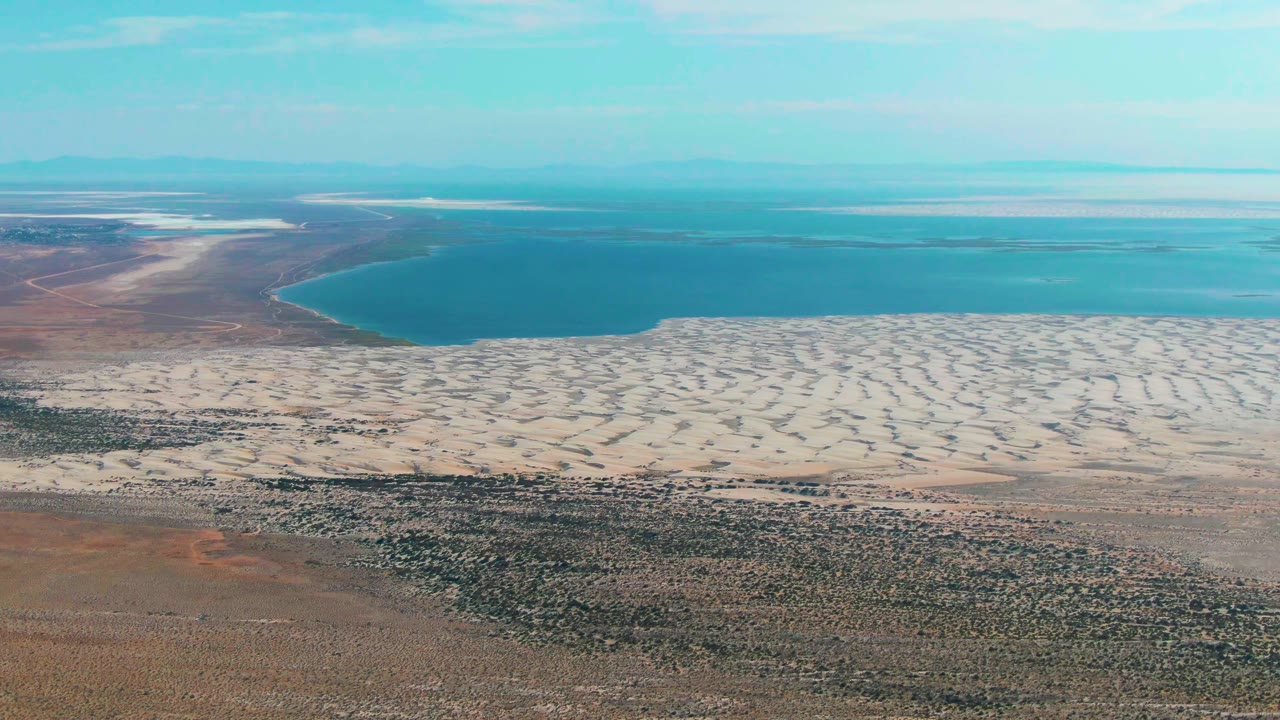Aerial frame over Guerrero Negro, Baja California Sur showing pale dune ridges beside a calm blue lake, with scrubby desert foreground, distant horizon, and no visible people.