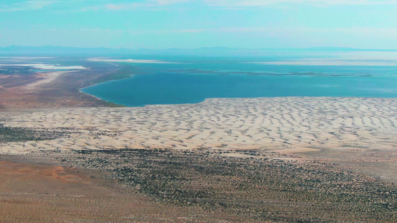 Aerial frame over Guerrero Negro, Baja California Sur with a curved lake edge, pale patterned dunes across the middle, scrubland below, hazy distance, and no visible people.
