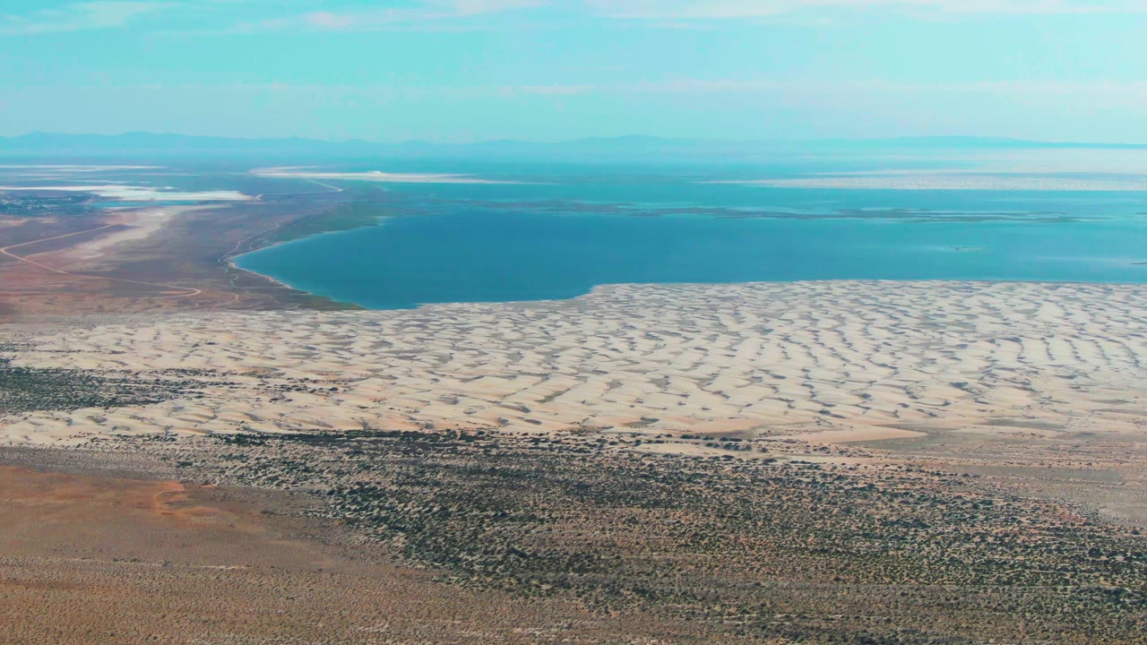 Aerial frame over Guerrero Negro, Baja California Sur showing a broad band of pale dune patterns below the blue lake, reddish desert flats at left, distant water bands, and no visible people.