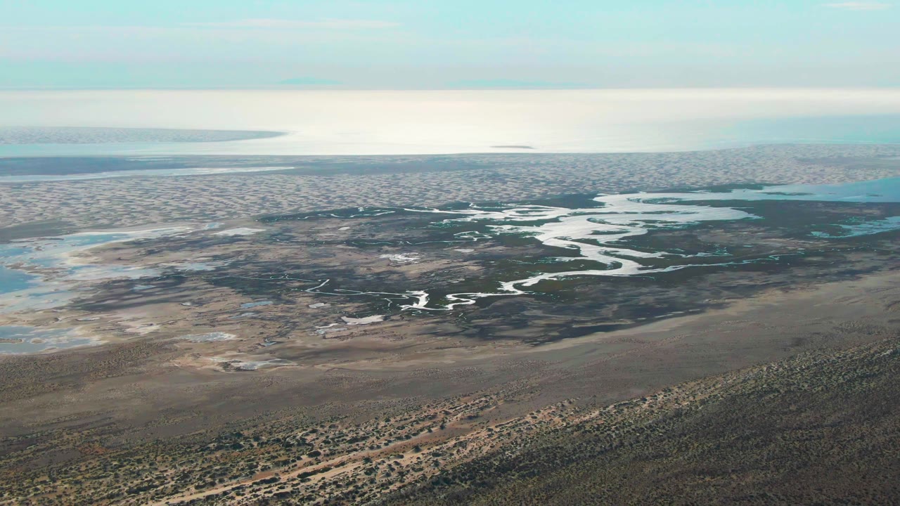 Aerial frame over Guerrero Negro, Baja California Sur, showing a pale winding river through dark wetlands, tan flats, blue coastal water, and no visible people.