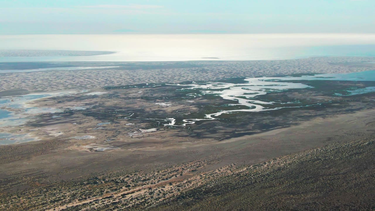 Aerial view in Guerrero Negro with the river channel centered across dark marshland, foreground scrubland below, pale flats to the left, and no visible people.