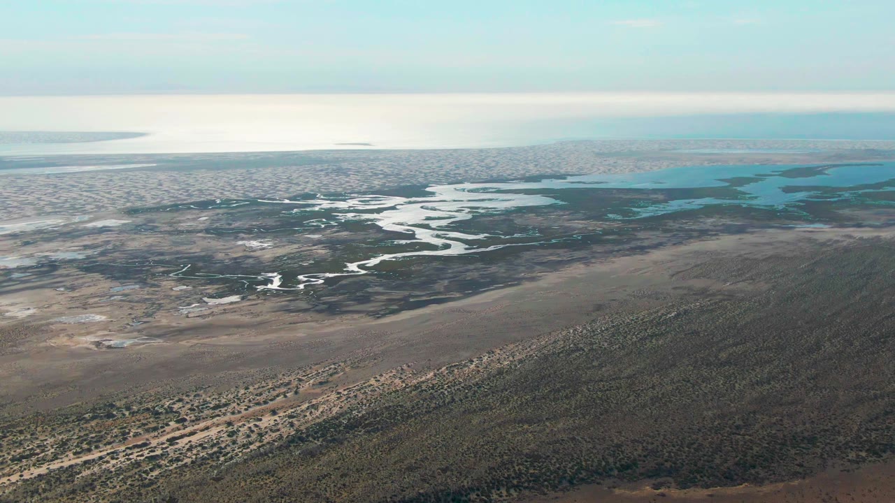 Opening aerial frame over Guerrero Negro, Baja California Sur, showing a winding wetland channel, sandy scrub terrain, distant blue-green water, and no visible people.