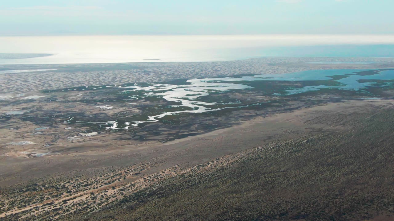 Aerial frame in Guerrero Negro, Baja California Sur, with bright meandering water cutting through darker marsh, dry scrubland in the foreground, and no visible people.
