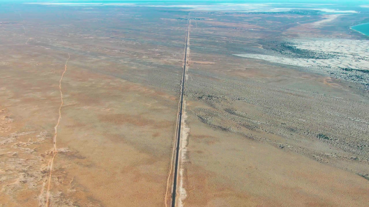 Aerial frame over Guerrero Negro, Baja California Sur, with a straight highway cutting through tan desert toward pale flats and blue water at the upper right, with no visible people.