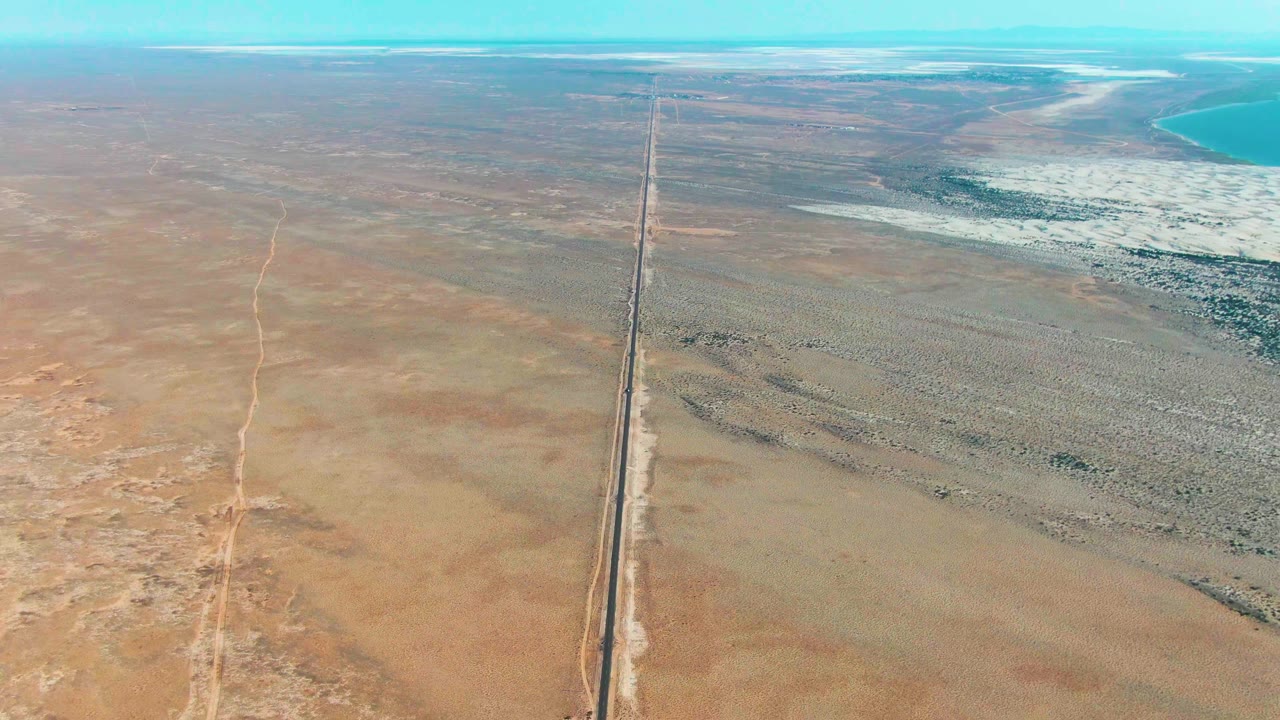 Aerial view in Guerrero Negro showing the highway centered through broad sandy desert, with pale flats to the right and a turquoise water edge near the horizon, with no visible people.