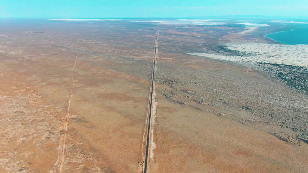 Drone-style aerial frame over Guerrero Negro with the straight road stretching from the lower center to the distant horizon, desert on both sides, and blue water visible at upper right, with no visible people.
