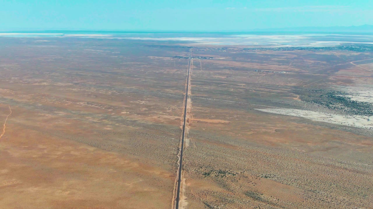 A later aerial frame over Guerrero Negro, Baja California Sur, where the straight road or rail line cuts through empty brown plains under a clear turquoise-blue sky with no visible people.