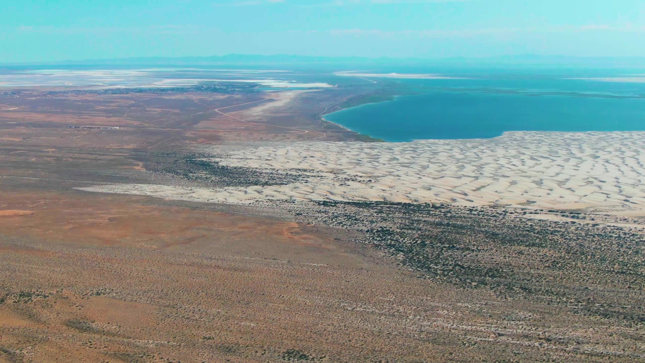 Aerial view of white salt flats beside blue water and brown desert terrain.