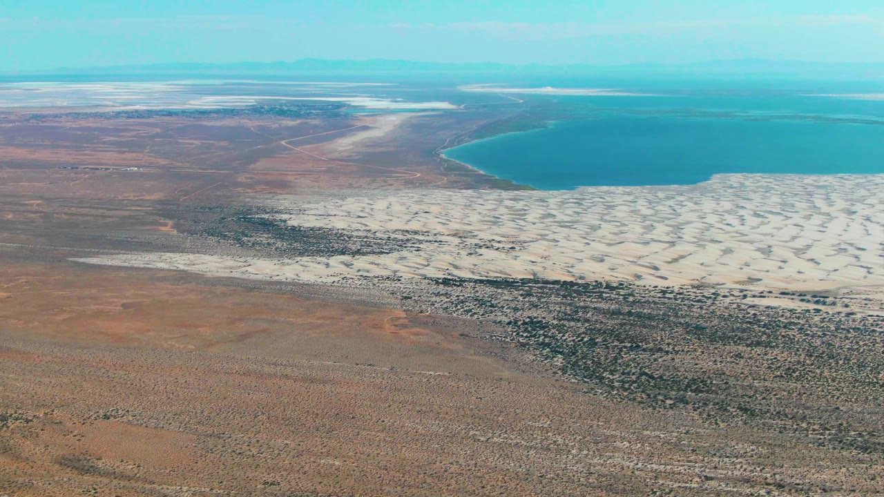 Second frame of the Guerrero Negro aerial view, with the white salt texture filling more of the midground below the blue water and no visible people.