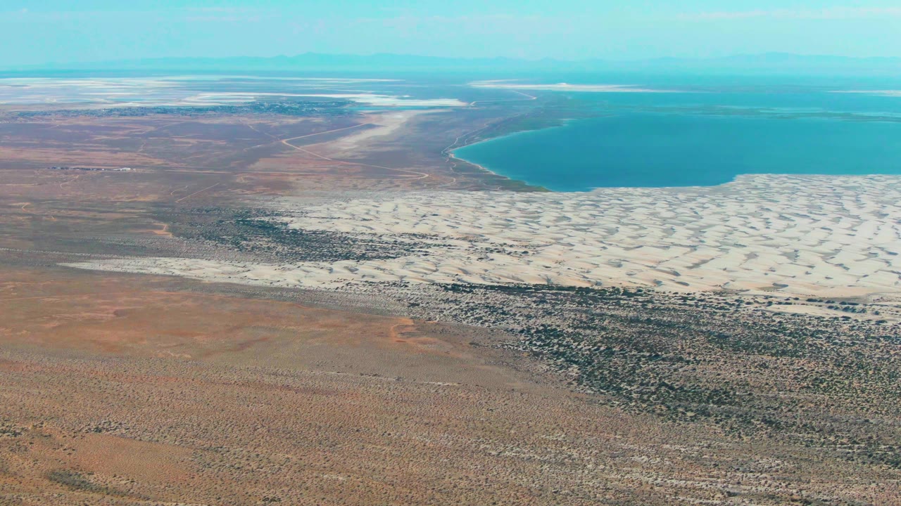 Third frame over Guerrero Negro showing the salt flats stretched below a curved blue water edge, with reddish brown arid ground in front and no visible people.