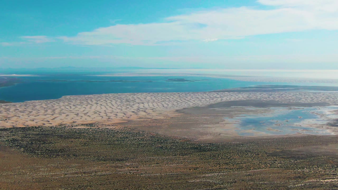Aerial view in Guerrero Negro, Baja California Sur, Mexico, showing deep blue ocean beyond a pale sandy shoreline, shallow tidal flats, brown-green land, bright sky, and no visible people.