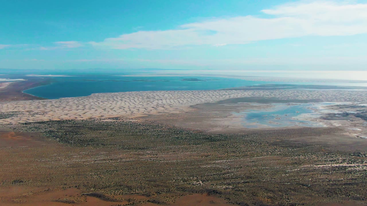Drone-height view in Guerrero Negro with a curved dark-blue water area at left, pale shoreline across the center, shallow turquoise pools to the right, inland terrain below, and no visible people.